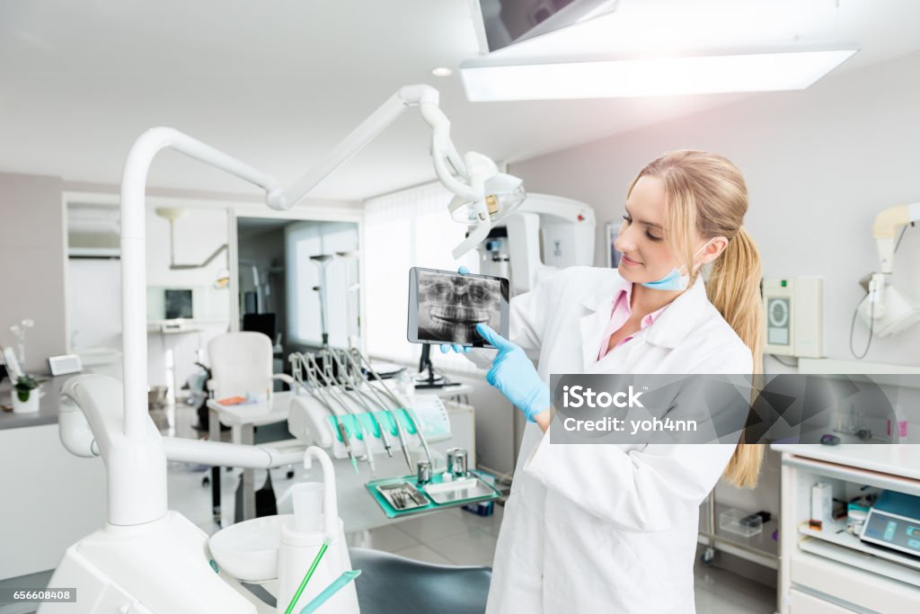 Showing the dental record on tablet Horizontal color image of female dentist using digital tablet and showing dental x-ray image. Young woman wearing white lab coat, surgical blue mask and gloves. Looking, examining and smiling. Computer Software Stock Photo