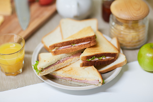 Close-up shot of appetizing breakfast prepared for family: sandwiches with ham and lettuce, apple, glass of orange juice, jar with cereals and cutting board located on table