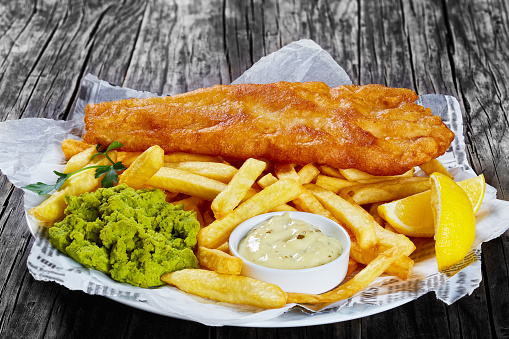 delicious crispy fish and chips - fried cod, french fries, lemon slices, tartar sauce and mushy peas on plate on paper, on wooden table, front view from above, close-up