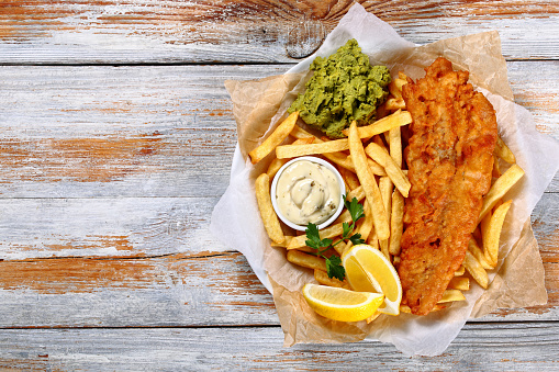 delicious crispy fish and chips - fried cod, french fries, lemon slices, tartar sauce and mashed peas on plate on paper on old wooden tabletop, authentic british recipe, view from above, blank space left