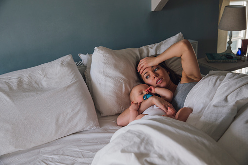 Young mother is lying in her bed with her baby daughter asleep in her arms. She has her hand on her head and looks very tired and stressed.