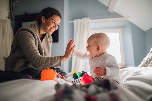 Baby girl is sitting on a double bed with her mother and they have baby toys and stacking cups around hem. The little girl is high-fiving her mother.