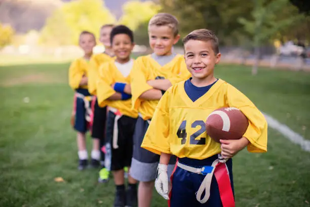 Five young boys dressed in flag football uniforms stand posing for a team portrait. The children are smiling and looking at the camera while standing on a football field in Utah, USA.