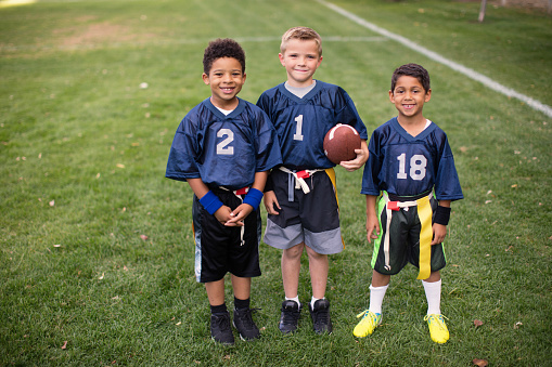 Three young boys dressed in flag football uniforms stand posing for a portrait. The children are smiling and looking at the camera while standing on a football field in Utah, USA.