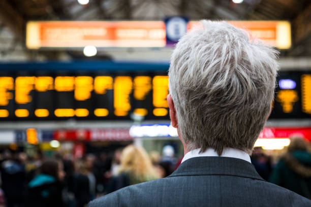Senior businessman waiting for train with departure boards in background, London, UK Rear view of a senior businessman, wearing suit and white shirt, waiting for his train. The orange writing on the digital departure screens are blurred out of focus in the background in a train station in London, UK. Also crowds of unrecognisable people are defocused behind the businessman, who has grey hair and is in his sixties, and of caucasian ethnicity. Close up colour image with copy space. back of head photos stock pictures, royalty-free photos & images