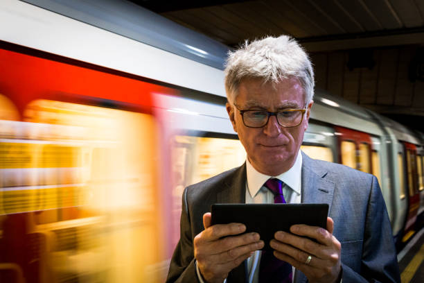 Senior businessman waiting for an underground train and looking at his digital tablet, London, UK A senior businessman in his 60s, wearing a grey suit with purple tie, waits for his train on the London underground, while he looks intently at his digital tablet. Behind him we can see the moetion bllur of an underground train, illuminated as it speeds past. Horizontal colour image with copy space. digital underground stock pictures, royalty-free photos & images