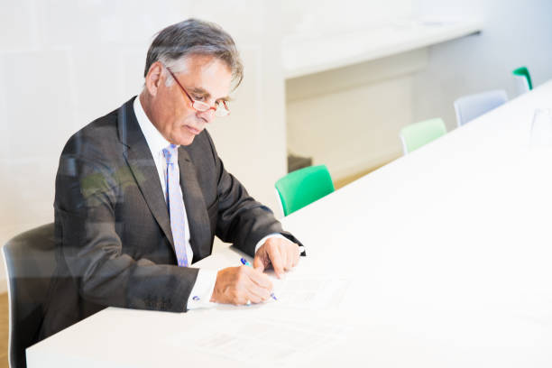 Senior businessman working late, sitting alone in modern meeting room signing and finalising the contracts. Shot through conference room glass window.
