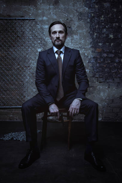 Handsome bearded man looking dangerous Super villain. Good looking brunette sharp gentleman wearing a classic suit and sitting on a chair in a dark room while holding a gun godfather godparent stock pictures, royalty-free photos & images