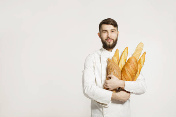istock Handsome smiling baker in uniform holding baguettes with bread shelves on the white background. Handsome man holding warm bread in his hands on white background. 656790652