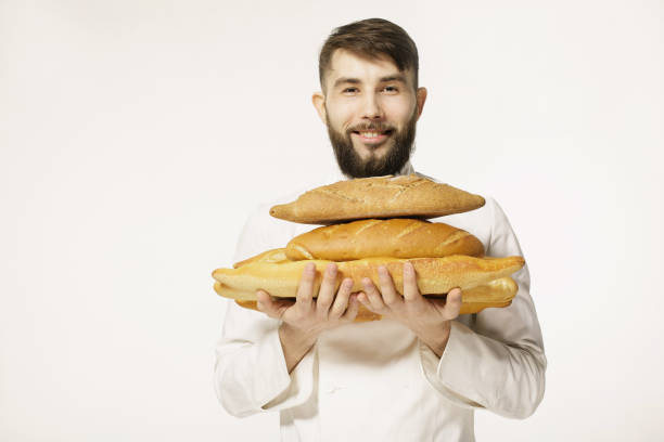 istock Handsome smiling baker in uniform holding baguettes with bread shelves on the white background. Handsome man holding warm bread in his hands on white background. 656790688
