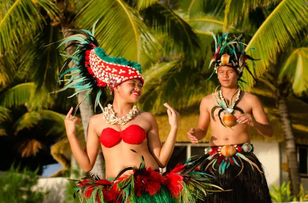 Portrait of attractive young Polynesian Pacific Island Tahitian male and female dancers in colorful costumes dancing on tropical beach during sunset with palm trees in the background.