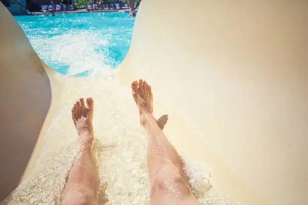 Point of view photo of a men going down a waterslide at an outdoor waterpark during a warm summer day. Lots of copy space. Focus on the feet and water slide