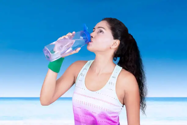 Portrait of beautiful woman drinking water on a bottle while standing at the beach