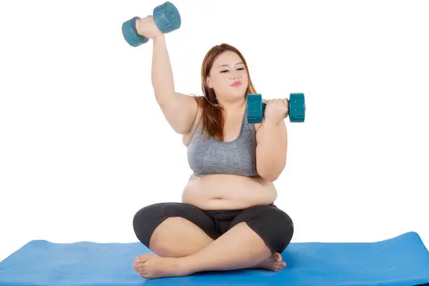 Portrait of blonde woman with overweight body, doing workout with two dumbbell while sitting on the mat