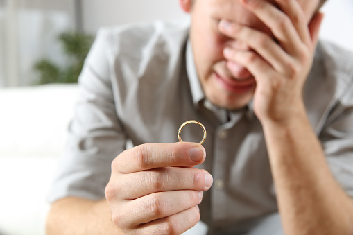Closeup of a sad husband lamenting after divorce holding the wedding ring in a house interior