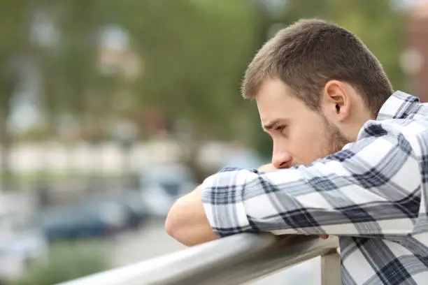 Side view portrait of a sad single man looking down from a balcony of a house  with an urban background