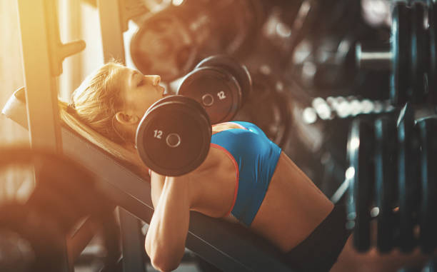 Gym workout. Closeup side view of a mid 20's blond woman doing dumbbell flying exercise on an incline bench. The sunlight is flaring from the left. She's wearing black short and blue tank top. Toned image. sportsman professional sport side view horizontal stock pictures, royalty-free photos & images