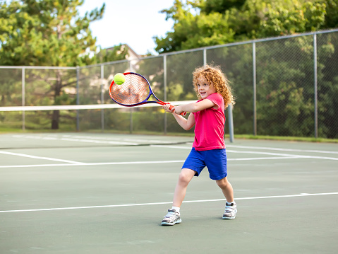 Young girl with curly red hair practicing hitting a tennis ball on an outdoor tennis court on a late summer/early autumn day. The girl is wearing a bright pink shirt with blue shorts. The tennis ball is just about to hit her racket, and the girl has a smile on her face.