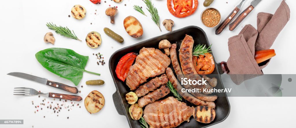 steak and sausage cooked on a grill with grilled vegetables in a cast iron pan on a white background steak and sausage cooked on a grill with grilled vegetables in a cast iron pan on a white background, top view. Flat lay Barbecue Grill Stock Photo