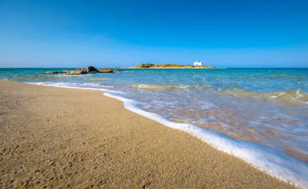 Photo of Typical summer image of an amazing pictorial view of a sandy beach with a boat on the beach and an old white church in a small island at the background, Malia, Crete, Greece.