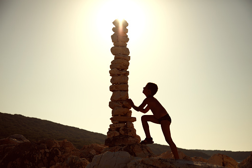 Little boy standing next to the huge stack of rocks and supporting their balance. Sun is positioned over top of the stack, so the boy and balanced stones sculpture looks like silhouette