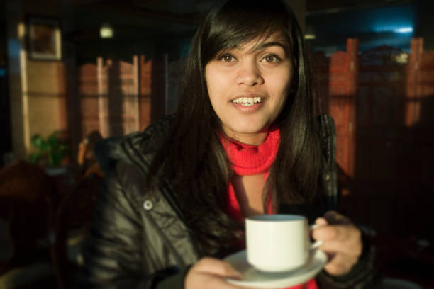 Indoor image of a young, happy Asian woman drinking coffee and giving toothy smile. One person, waist up, horizontal composition with copy space and selective focus.