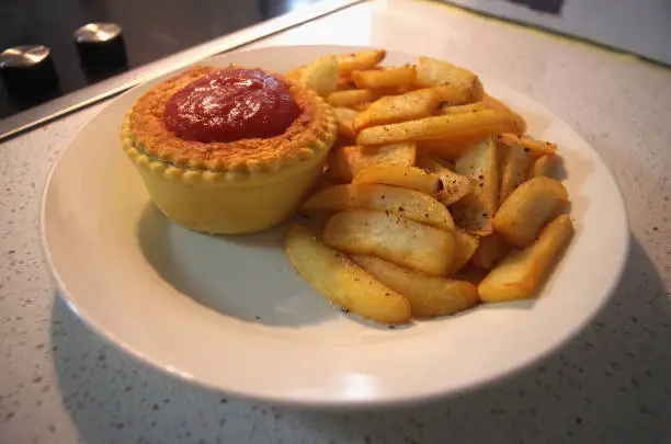 A Meat Pie with Tomato Sauce, served with Golden Hot Chips on a White Plate near a Stovetop on a Kitchen Counter.