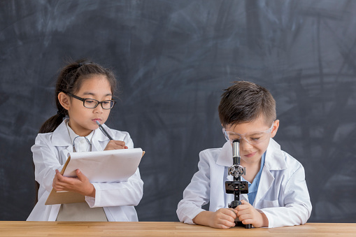 Elementary school students record finding while looking into a microscope. The male student uses the microscope while the female student records observations on a note pad.