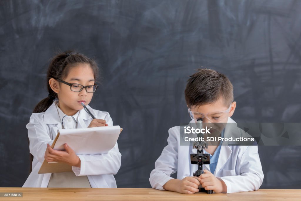 Smart school children use microscope Elementary school students record finding while looking into a microscope. The male student uses the microscope while the female student records observations on a note pad. Asian and Indian Ethnicities Stock Photo