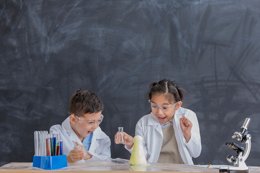 Elementary age Asian brother and sister watch for a chemical reaction while conducting chemistry experiment. They are wearing safety glasses and lab coats.