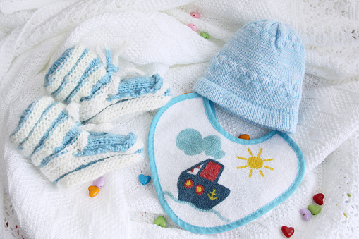 blue knitted newborn baby booties, hat and bib on crocheted blanket white background with colorful hearts with copy space