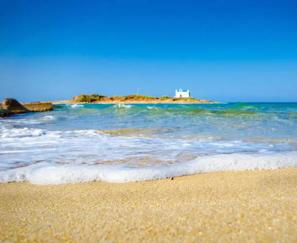 Photo of Typical summer image of an amazing pictorial view of a sandy beach with a boat on the beach and an old white church in a small island at the background, Malia, Crete, Greece.