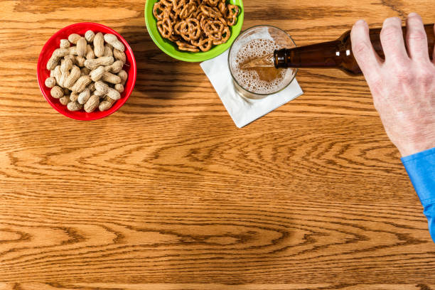 An overhead view of a middle-aged Caucasian businessman pouring his beer from a brown bottle into a beer glass. The bar is made out of oak, it has a bowl of whole peanuts and a bowl of pretzels.