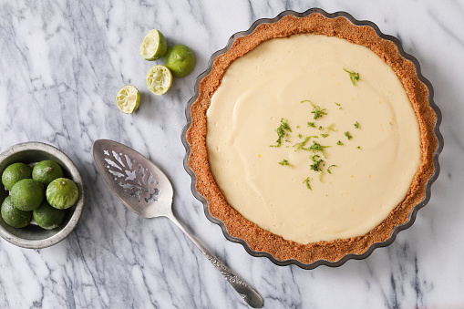 An overhead close up, horizontal photograph of a fresh, home made key lime pie garnished with lime zest, a bowl of fresh limes and several squeezed ones and a metal serving spatula can be seen next to the pastry.
