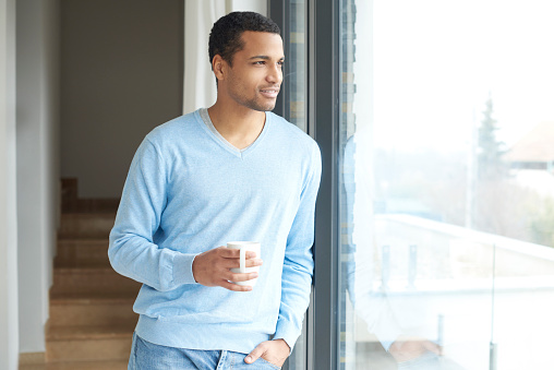 Shot of a handsome man looking pensively out the window while drinking coffee in her modern home.