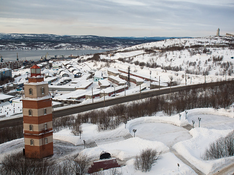 City of Murmansk and the Memorial Complex of the sailors who died in peacetime with beacon tower against the backdrop of the sea port and snow covered streets