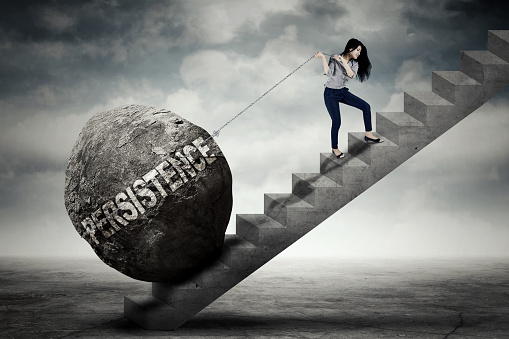 Young female entrepreneur is pulling big stone with persistence word while climbing on the stair