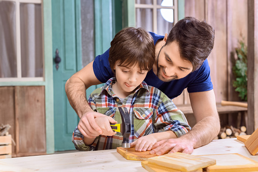 portrait of father teaching concentrated son how to hammering nail in wooden plank