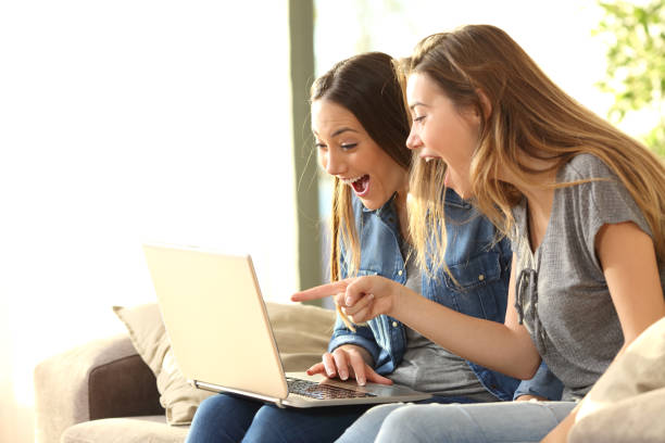 Excited roommates on line at home Two excited roommates reading good news on line with a laptop sitting on a couch in the living room at home with a window in the background excitement laptop stock pictures, royalty-free photos & images