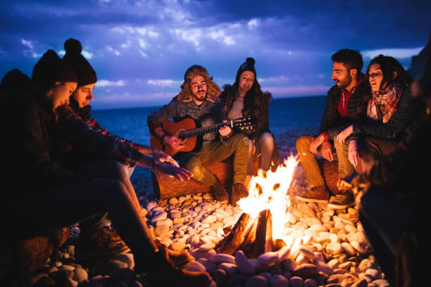 Friends playing guitar and singing around bonfire at the beach Young hipster friends with a guitar singing around a campfire at a beach at dusk bonfire stock pictures, royalty-free photos & images
