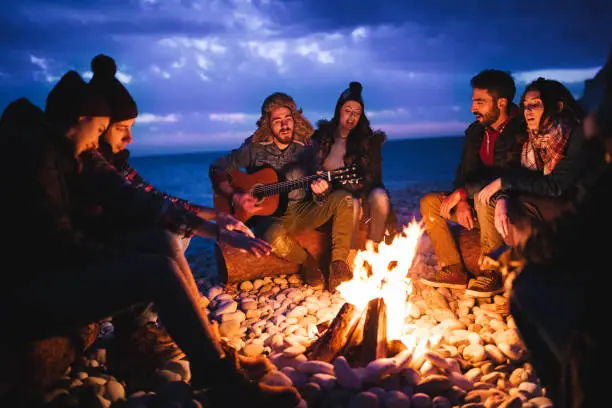 Young hipster friends with a guitar singing around a campfire at a beach at dusk