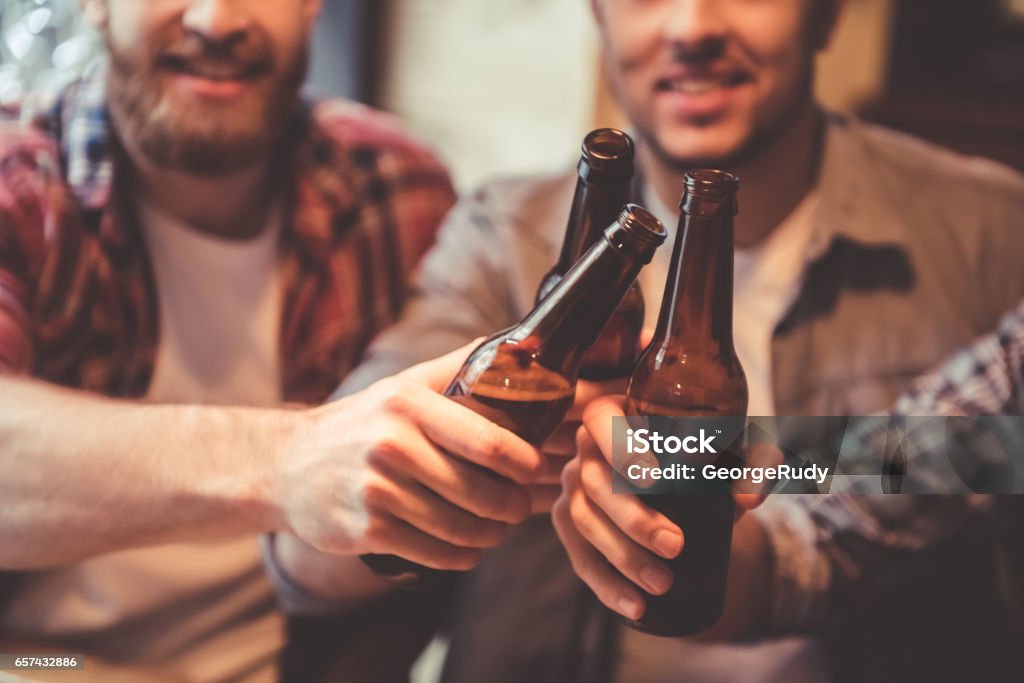 Friends at the pub Cropped image of handsome friends clinking bottles of beer and smiling while resting at the pub Beer - Alcohol Stock Photo