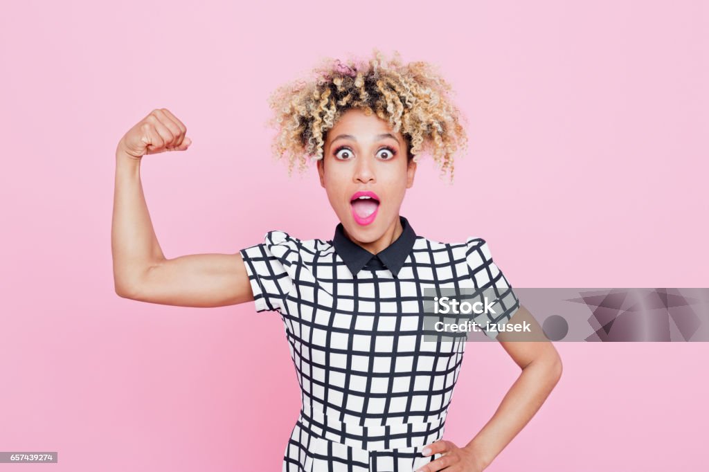 Excited afro american young woman flexing muscle Studio portrait of excited afro american young woman flexing muscle. Pink background. Bicep Stock Photo