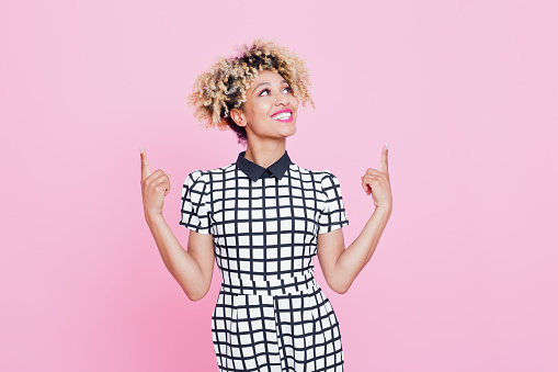 Studio portrait of happy afro american young woman pointing with index fingers at copy space. Pink background.