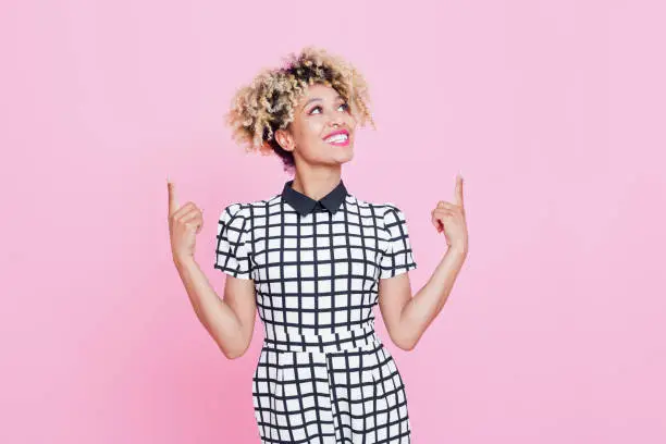 Studio portrait of happy afro american young woman pointing with index fingers at copy space. Pink background.