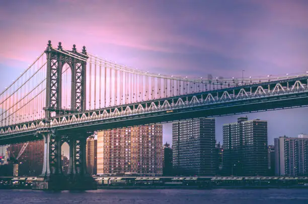The world famous Manhattan Bridge in New York City at sunset with a dramatic purple cloudscape and the island of Manhattan in the background with the east river in the foreground. One of the most famous tourist destinations in the world with a retro styled look to the photo.