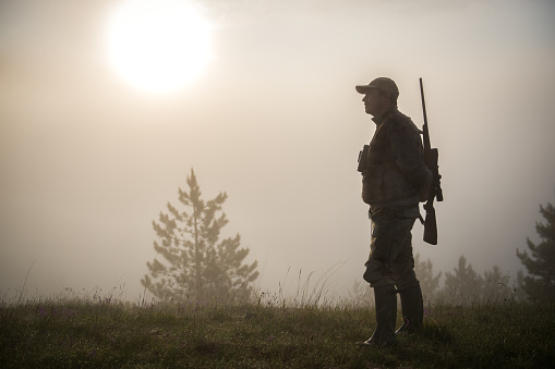 Portrait of hunter with rifle and binoculars in the forest. Bright sunlight is breaching through the fog in the background.