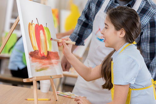 Cute Hispanic preteen girl enjoys painting in art class. An art teacher is helping her.