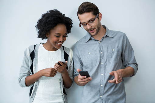 Man and woman standing against white wall and using mobile phone in office