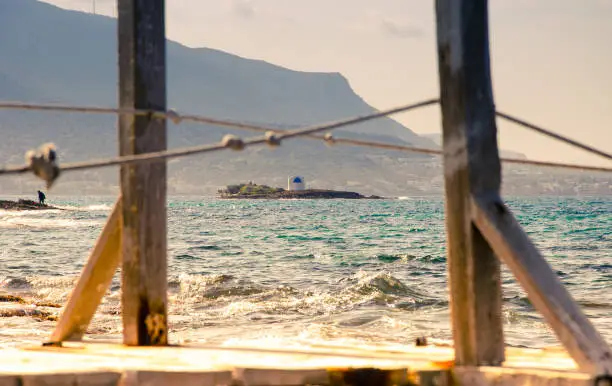 Photo of Typical summer image of an amazing pictorial view through a frame of a wooden pier and rops of an old white church in a small island, Malia, Crete, Greece.